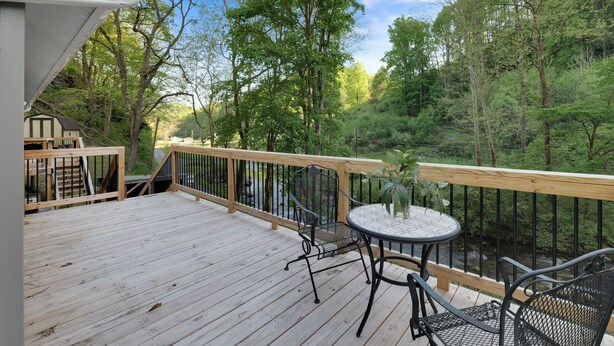 A wooden deck with a table and chairs, overlooking a forested area and a stream.