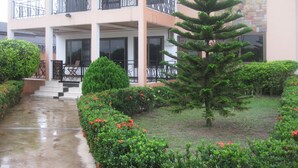 Um edifício com vários andares e varandas, um jardim bem cuidado com diversos arbustos e uma grande árvore conífera, e um caminho de concreto que leva à entrada.