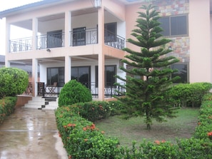 A multi-story building with balconies, a well-maintained garden with various shrubs and a large coniferous tree, and a concrete pathway leading to the entrance. - Room-ghouse Room B Ideal For Friends fam (Adenta)