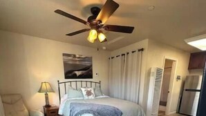 Ein Schlafzimmer mit Bett, Schreibtisch, Stuhl, Deckenventilator und einem Fenster mit Vorhängen.