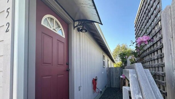 Ein schmaler Holzweg führt zu einer roten Tür mit einem kleinen Fenster darüber. Rechts sind ein Holzzaun und eine Holzbank zu sehen.