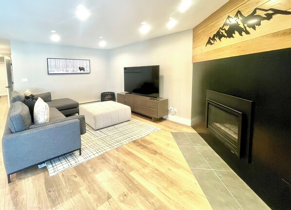 A modern living room with a grey sofa, a white ottoman, a flat-screen TV, a wooden accent wall, and a fireplace.