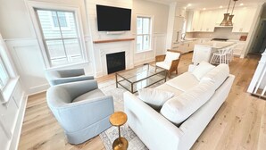 A spacious living room with a large white sofa, a grey armchair, and a glass coffee table. A flat-screen TV is mounted above a fireplace. The room features large windows and a ceiling fan.