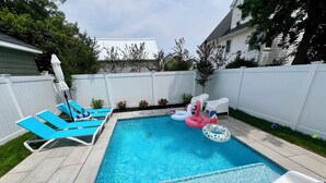 A rectangular swimming pool with clear blue water, surrounded by a white fence. There are lounge chairs and inflatable pool toys on the poolside.