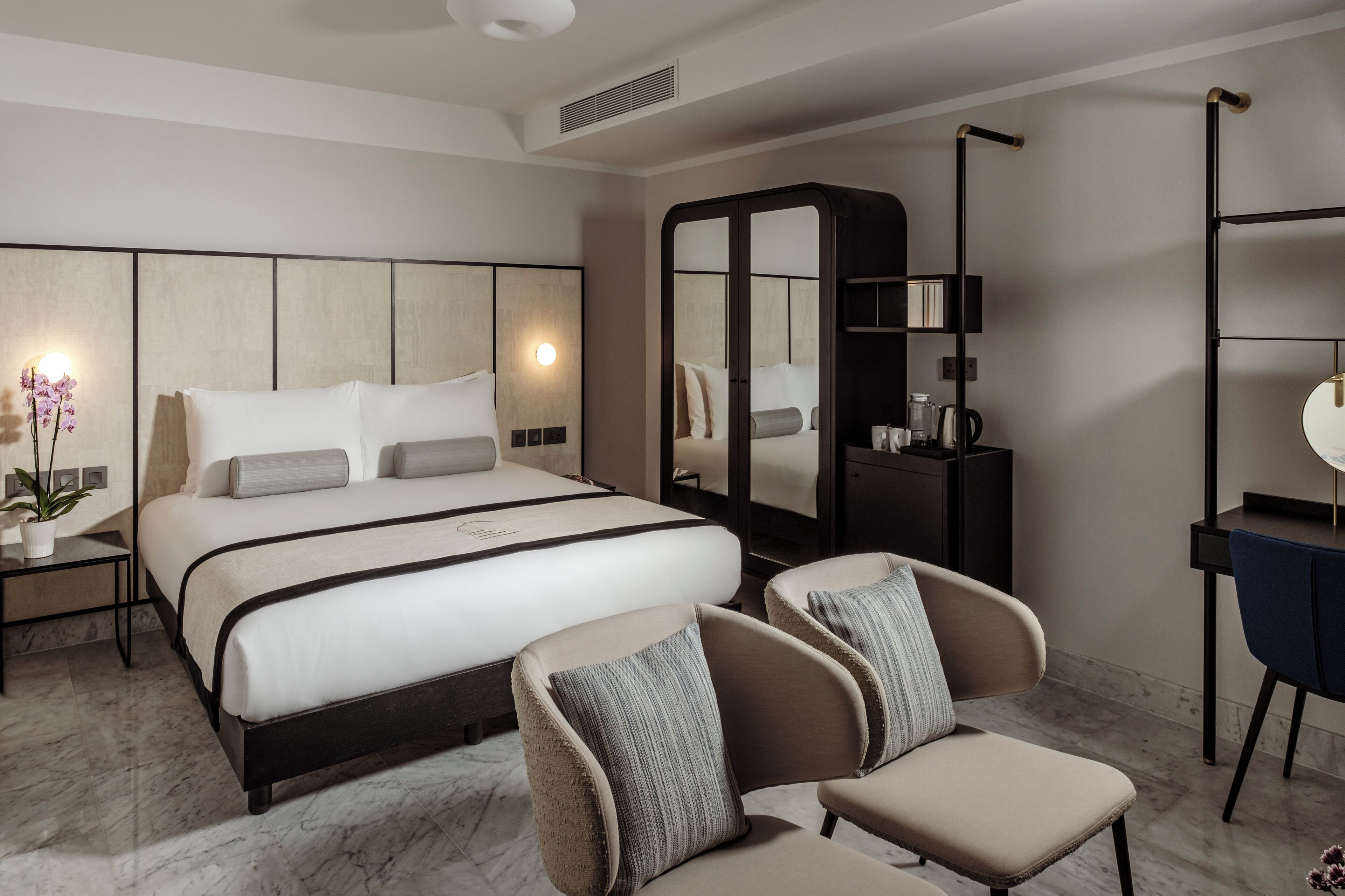 A modern hotel room with a large bed, a seating area with two chairs, a desk with a chair, a wardrobe, and a mirror.