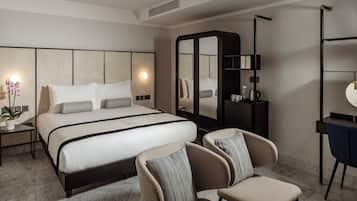 A modern hotel room with a large bed, a seating area with two chairs, a desk with a chair, a wardrobe, and a mirror.