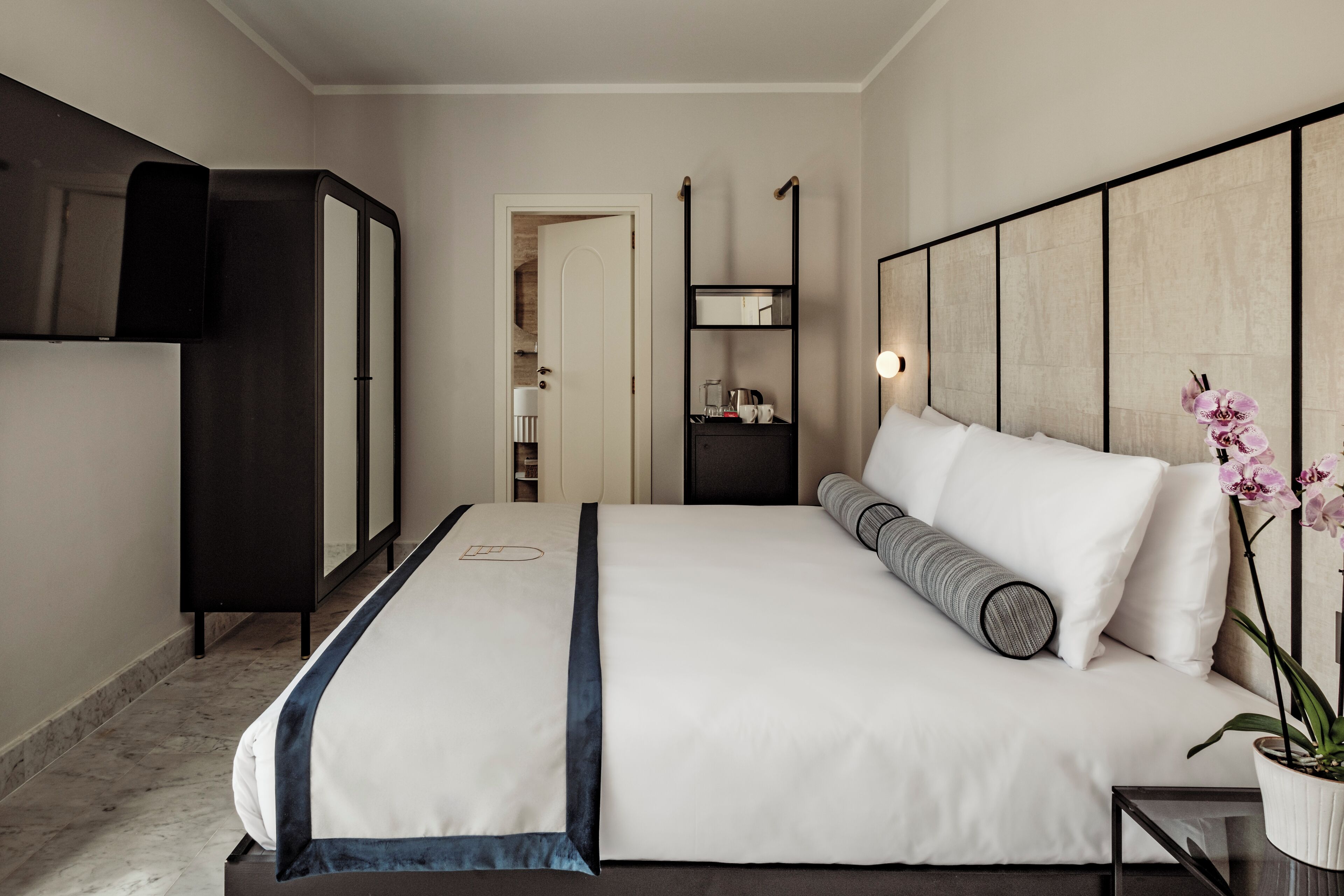 Moderni hotellihuone, jossa on suuri sänky, vaatekaappi, yöpöytä ja ruukussa oleva kasvi.