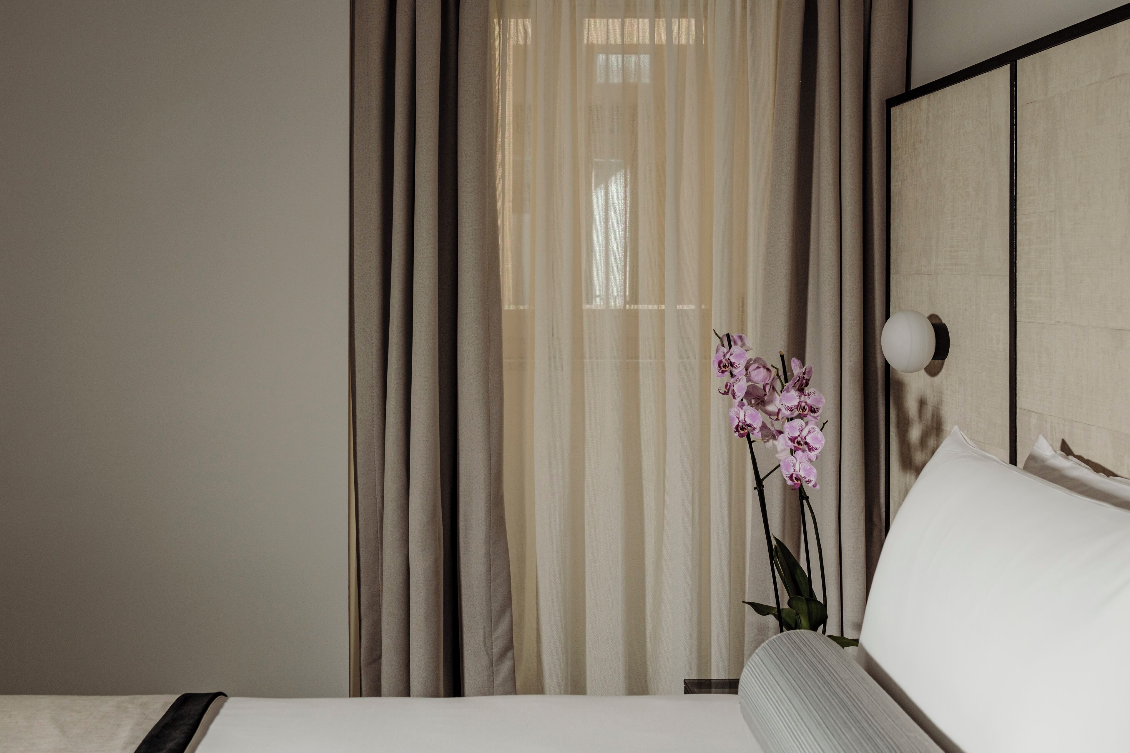 Habitació d'hotel amb un llit, una petita taula de nit, una làmpada i una finestra amb cortines translúcides.
