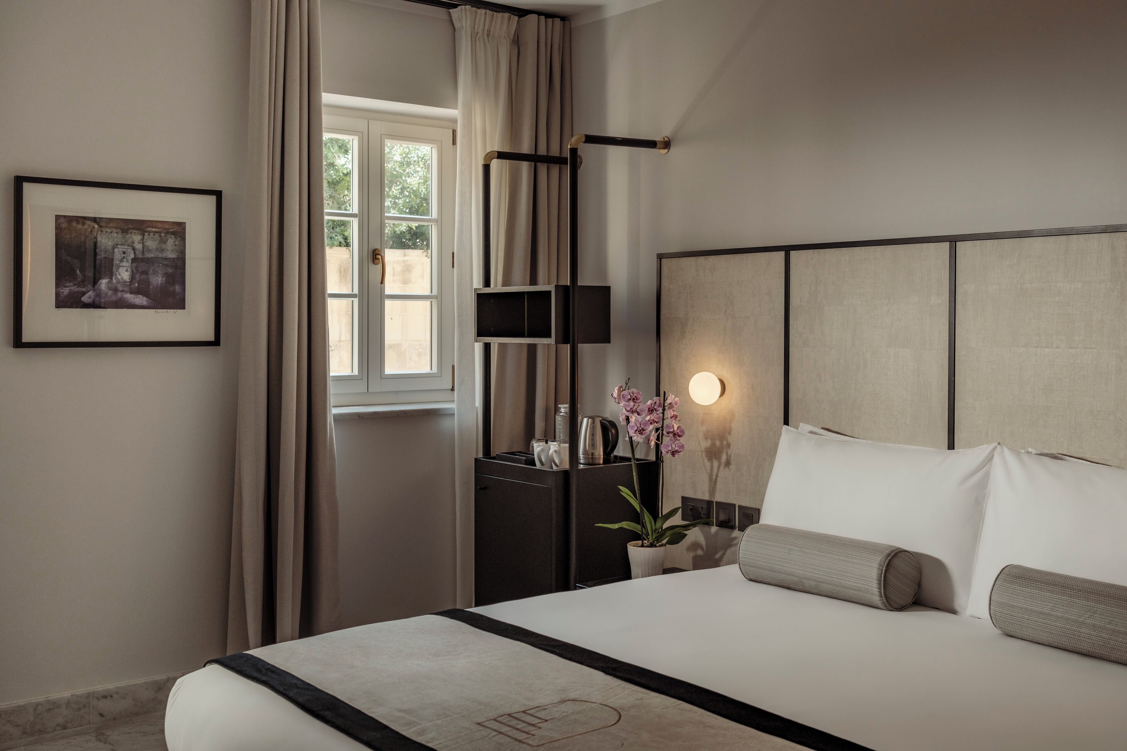 Ein Hotelzimmer mit Bett, Nachttisch, Lampe, einem gerahmten Bild und einem Fenster mit Vorhängen.