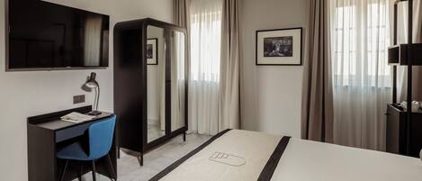 A hotel room with a bed, a desk with a chair, a TV, a wardrobe, and a framed picture on the wall.