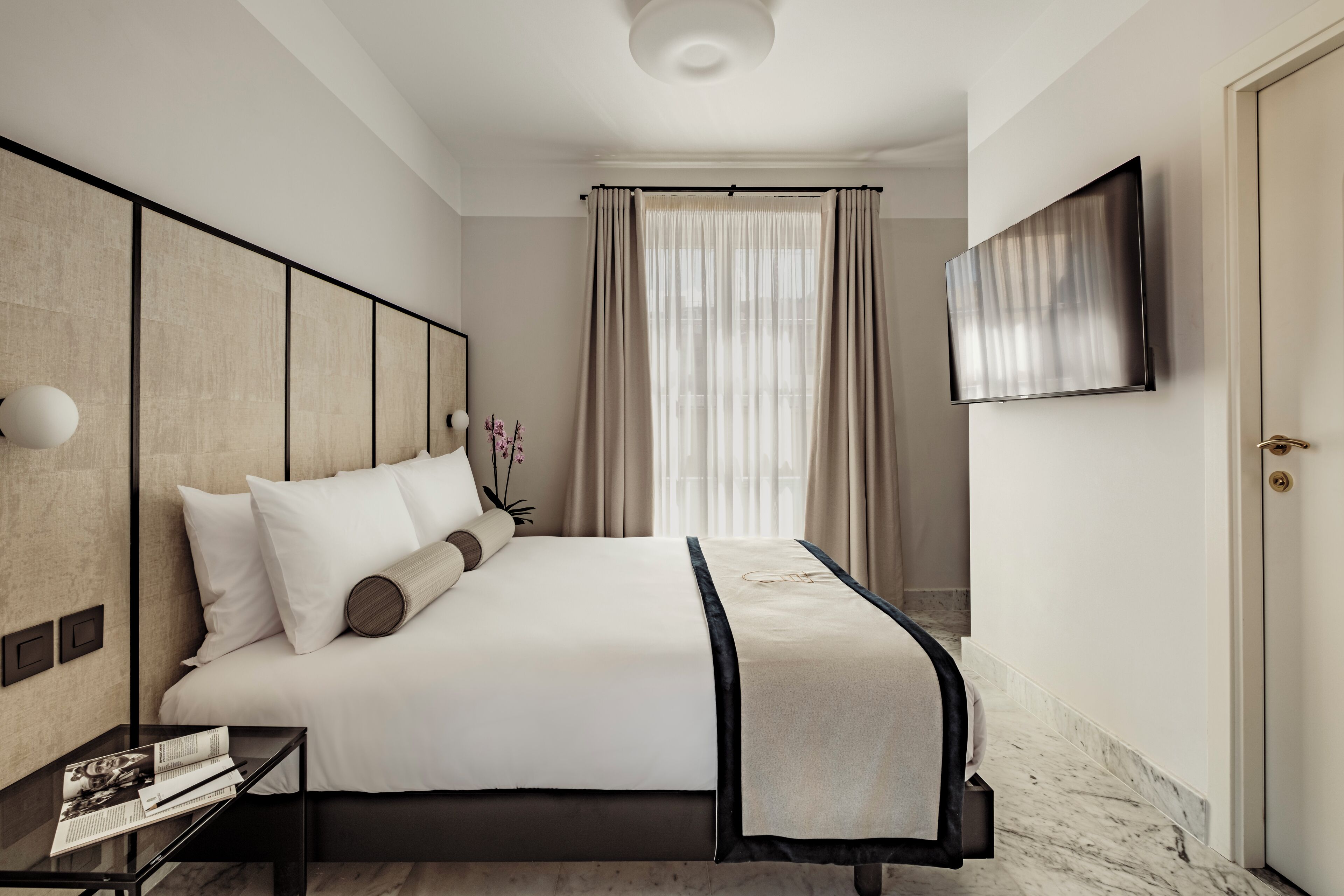 A modern hotel room with a large bed, a flat-screen TV mounted on the wall, and a window with curtains.