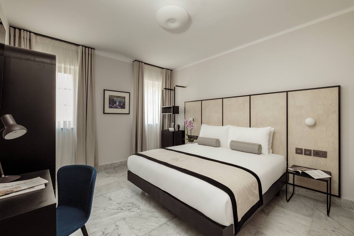 Una habitación de hotel moderna con una cama grande, un escritorio con lámpara, una silla y una mesita de noche con un libro y otra lámpara.