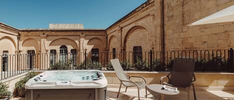 A hot tub on a patio with two chairs and a table, surrounded by a stone building with arches.