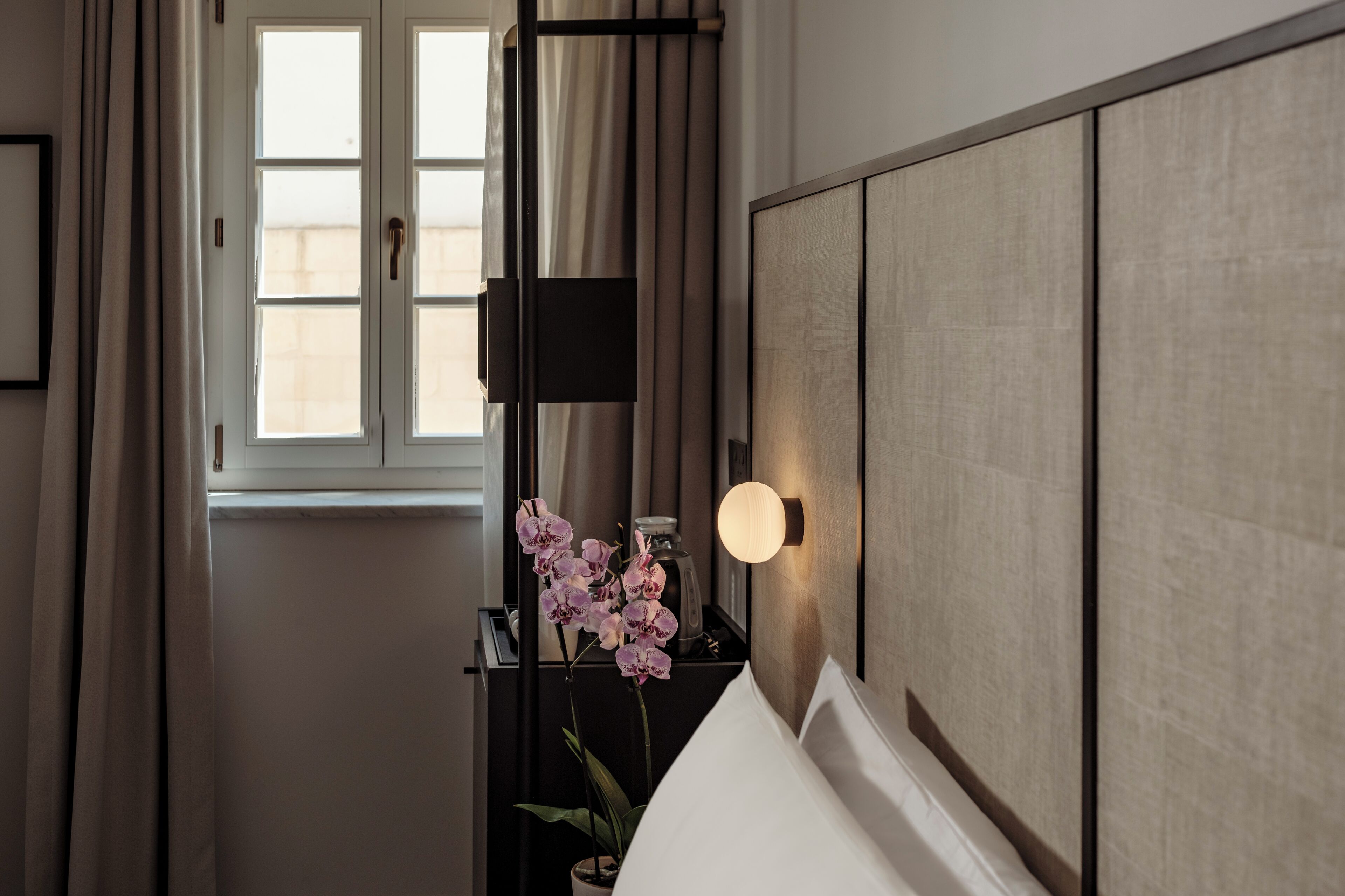 Una habitación con una cama, una mesita de noche con lámpara, una ventana con cortinas y una planta de orquídea en maceta.