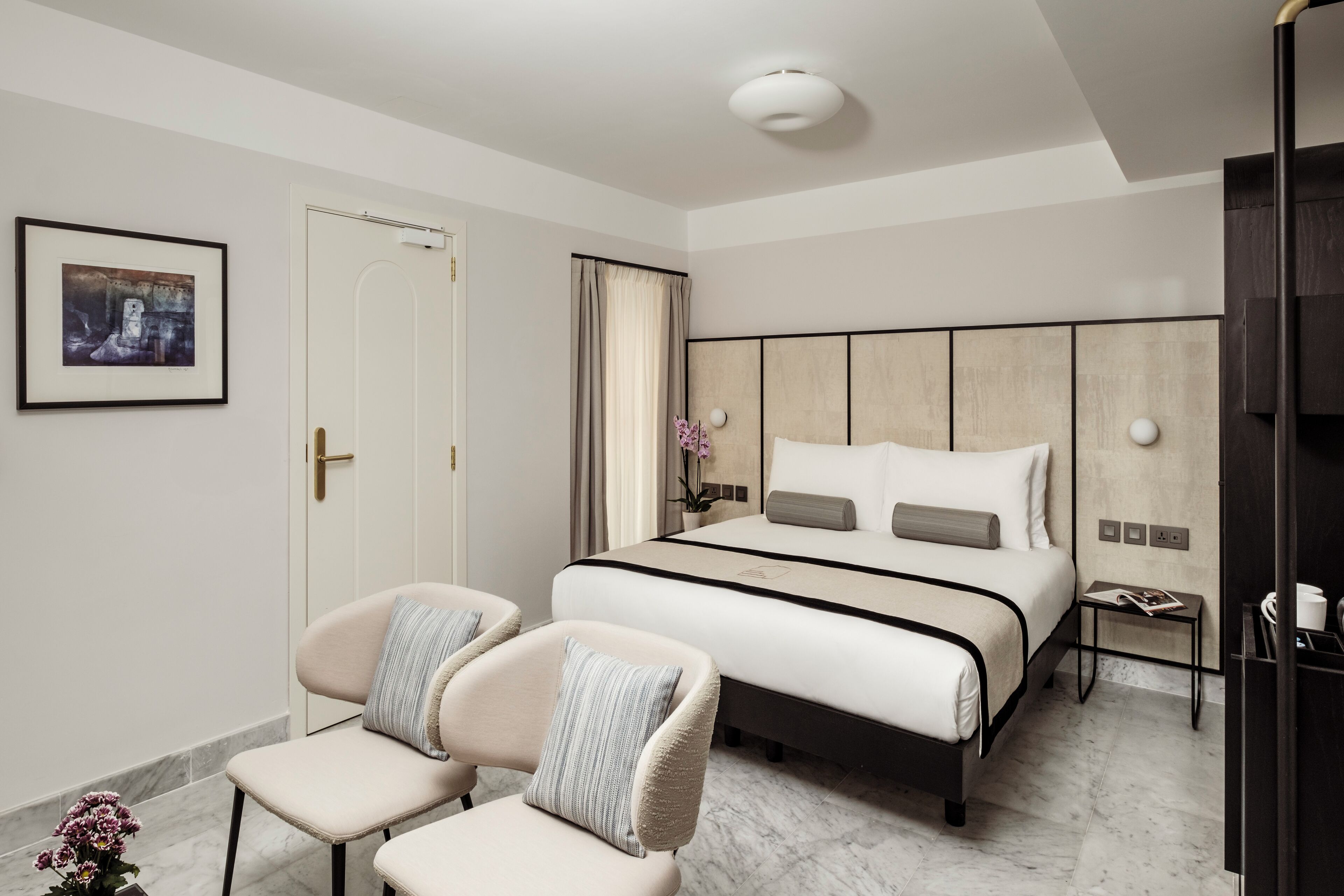 Ein modernes Schlafzimmer mit einem großen Bett, zwei Sesseln, einem kleinen Nachttisch und einem gerahmten Bild an der Wand.
