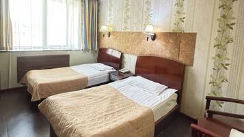 A hotel room with two beds, a wooden headboard, a desk, a chair, and a window with curtains.