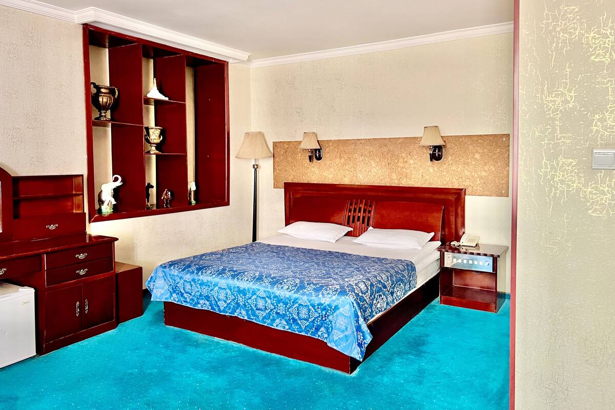 A hotel room with a large bed, a wooden dresser, a vanity with a mirror, and a small table with a lamp.