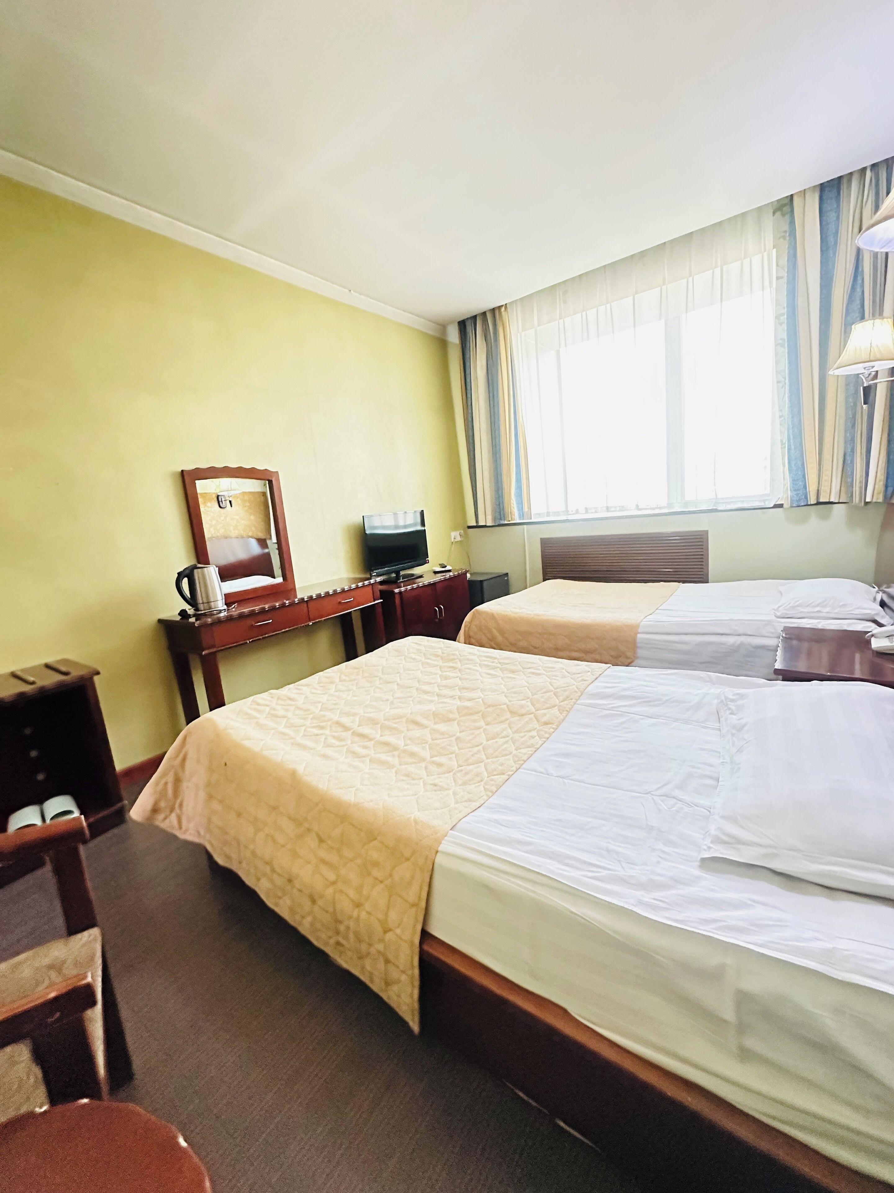A hotel room with two beds, a desk with a television, a mirror, and a window with curtains.