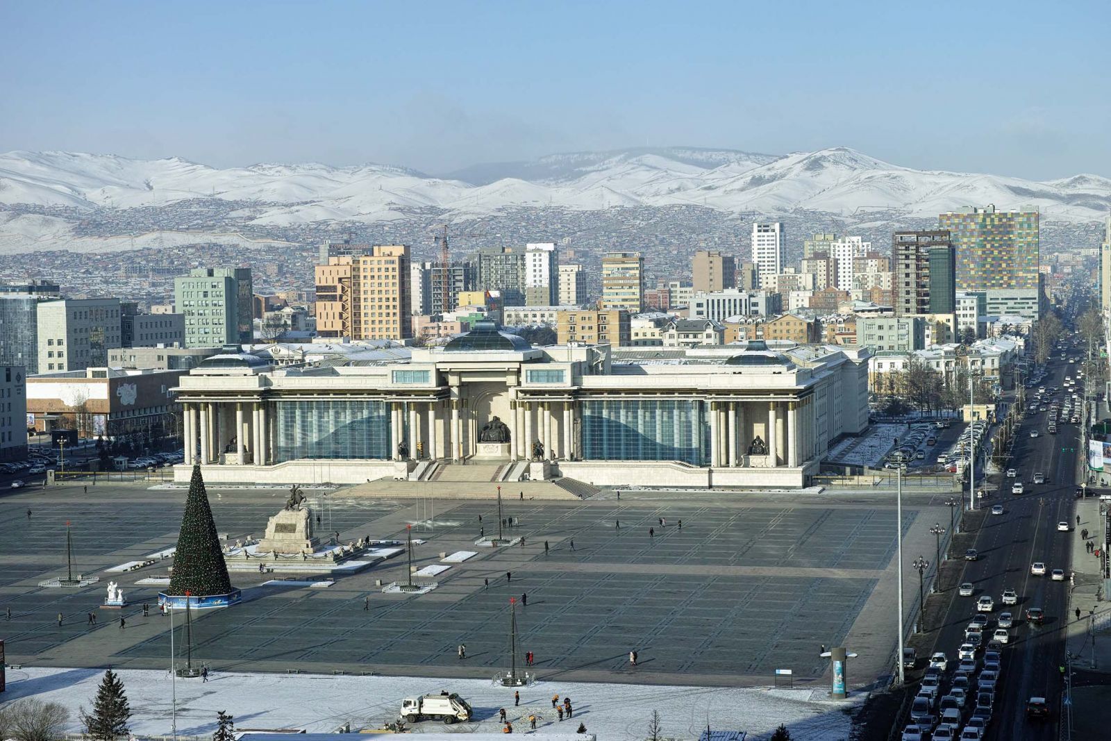 A large, open plaza with a prominent building featuring classical architecture, a large Christmas tree, and a snow-covered mountain range in the background.