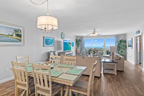 Ett rymligt vardagsrum med ett matbord, stolar och en soffa. Ett stort fönster ger utsikt över havet.