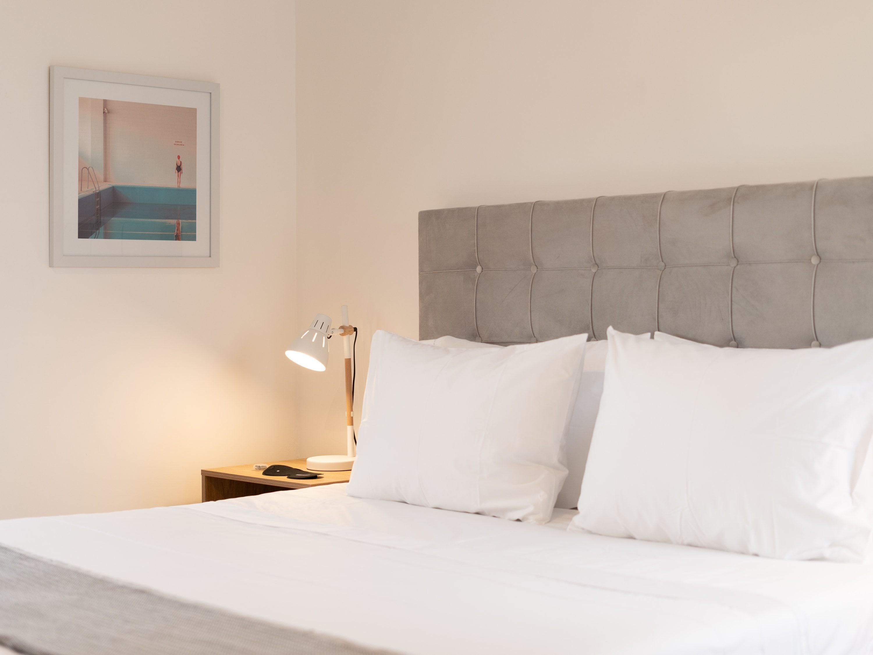 A neatly made bed with white linens, a grey tufted headboard, and a bedside table with a lamp and a phone.