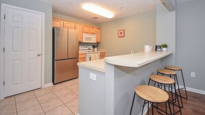 A kitchen with a refrigerator, microwave, and wooden cabinets. There is a small island with stools and a wall-mounted microwave.