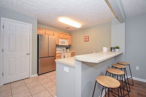Cocina con refrigerador, microondas y gabinetes de madera. Hay una pequeña isla con taburetes y un microondas instalado en la pared.