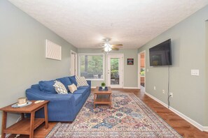 A living room with a blue sofa, a patterned rug, a ceiling fan, and a television on the wall.