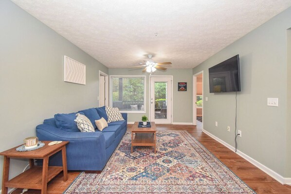 A living room with a blue sofa, a patterned rug, a ceiling fan, and a television on the wall.
