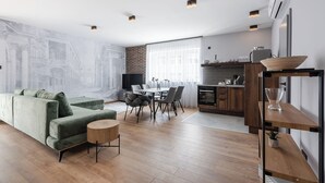 A modern living room with a green sofa, dining table, and chairs. There is a kitchen area with a refrigerator and a wooden shelf. - Radnoti Home (Veszprem)
