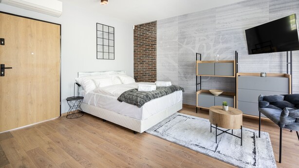 A modern bedroom with a wooden floor, a bed with white bedding, a brick accent wall, a mounted TV, and a small round wooden side table. - Radnoti Home (Veszprem)