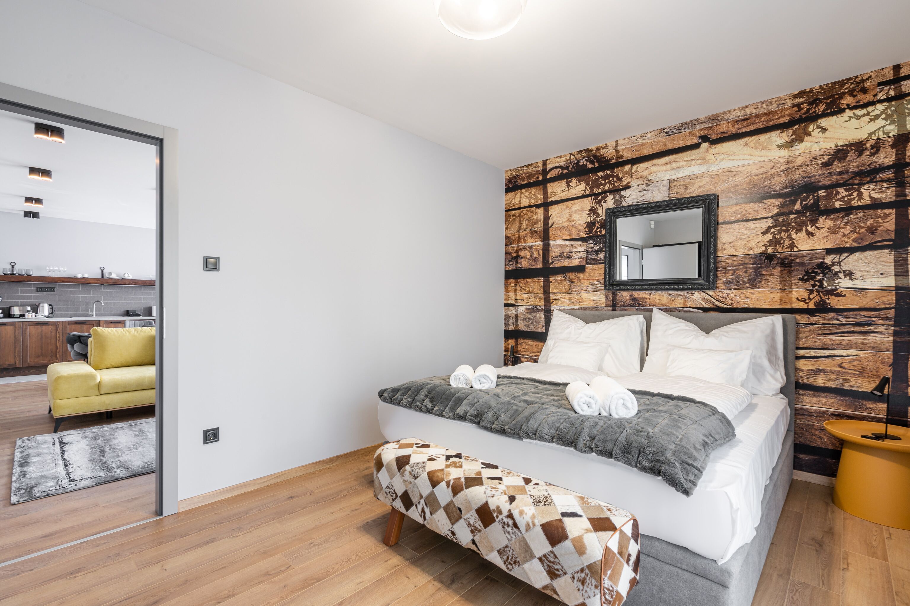 Een moderne slaapkamer met een groot bed, een houten accentmuur, een klein nachtkastje en uitzicht op de keuken.