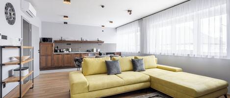 Una sala de estar moderna con un sofá seccional amarillo, piso de madera y una cocina al fondo.