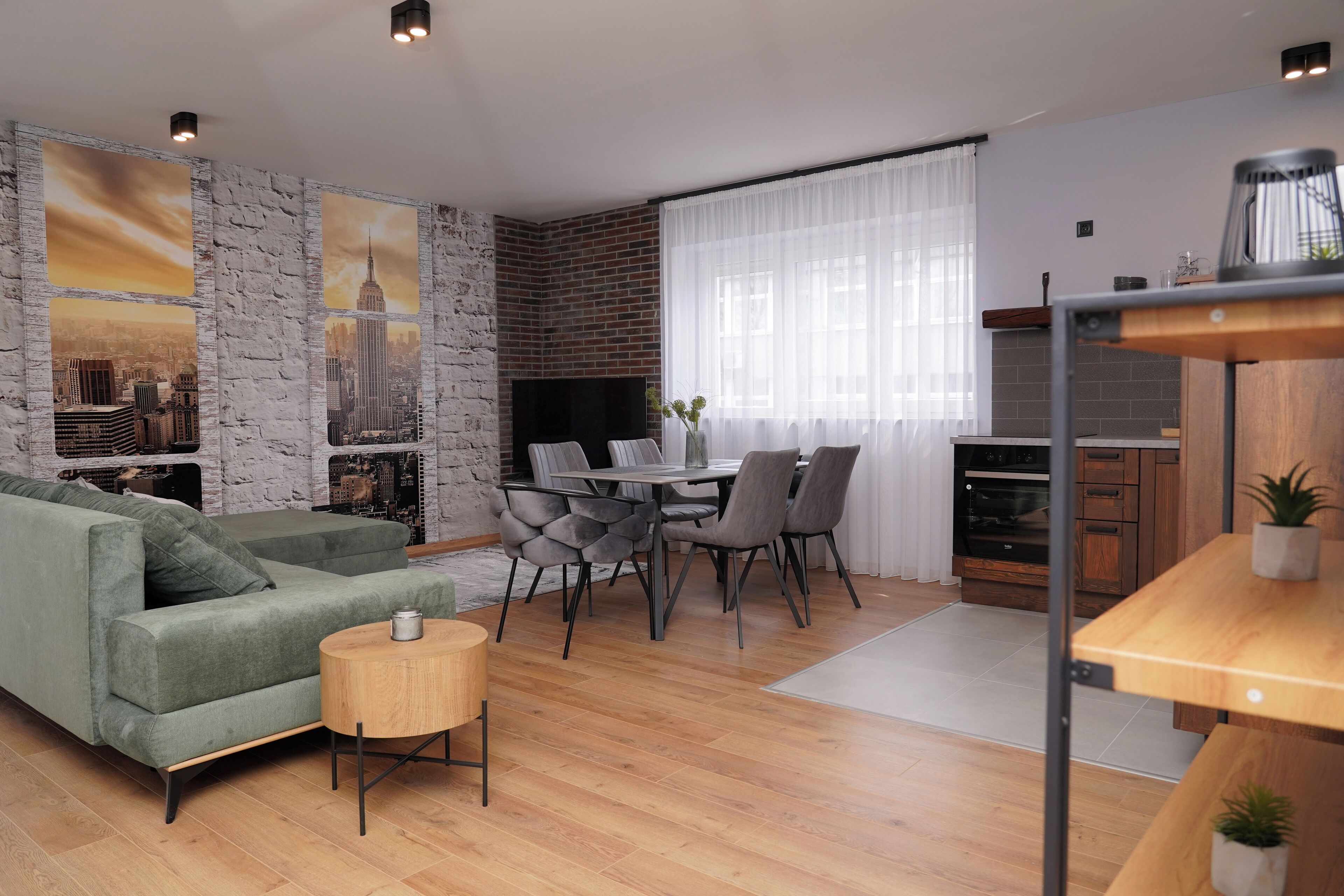 A modern living room with a green sofa, dining table, and chairs. There is a fireplace, a TV, and a kitchen area with a wooden table and cabinets.