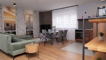A modern living room with a green sofa, dining table, and chairs. There is a fireplace, a TV, and a kitchen area with a wooden table and cabinets.