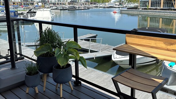 Eine Uferterrasse mit Kübelpflanzen, einem Holztisch und Blick auf Boote und Gebäude am Wasser.