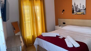 A hotel room with a bed, a nightstand, an air conditioning unit, a wall-mounted light, and a painting of a car.