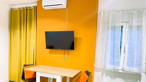 A room with a wooden table, two chairs, a mounted TV, an air conditioning unit, and a window with sheer curtains.
