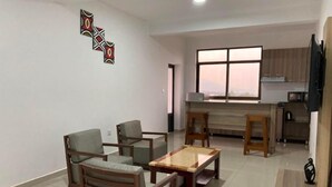 A modern living room with a sofa set, a coffee table, and a kitchen area with a window. - Rebero View Apartments (Kigali)
