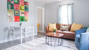 Un salon avec un canapé marron, un fauteuil bleu, une table à manger blanche avec des chaises, et un tableau d’affichage coloré accroché au mur.