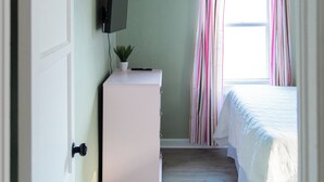 Une chambre à coucher avec un lit, un téléviseur fixé au mur, une petite commode blanche et une fenêtre avec des rideaux.