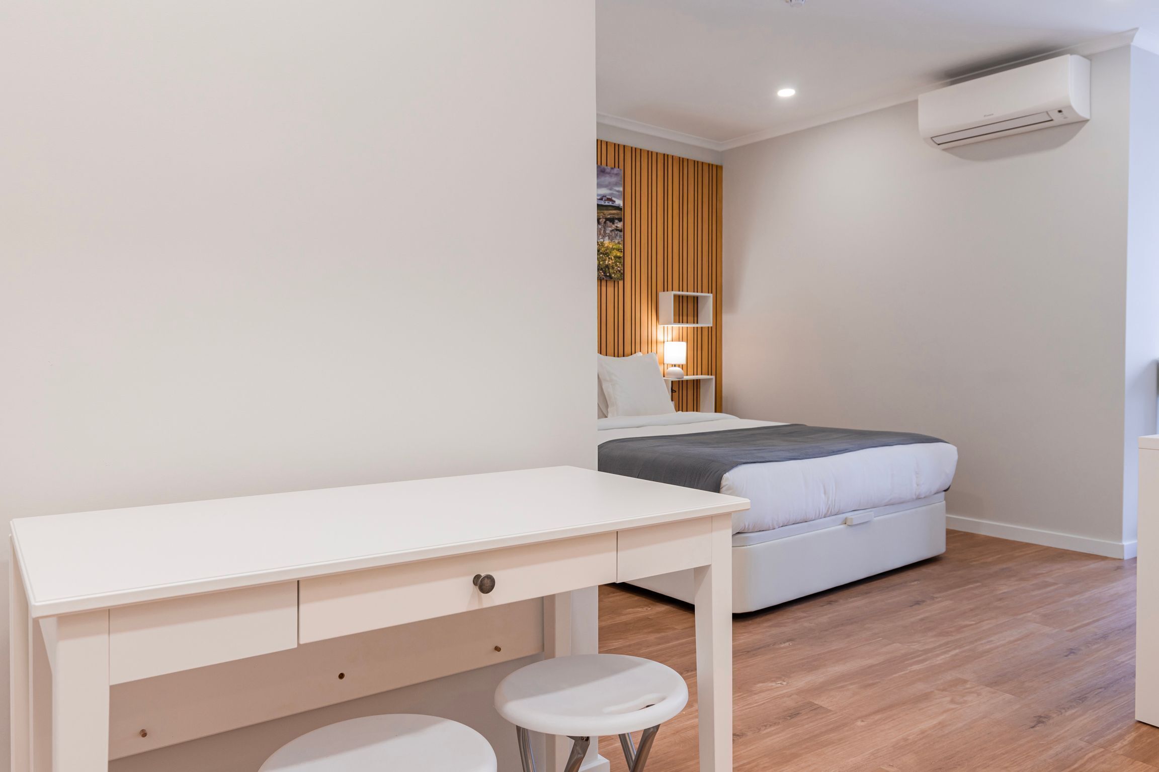 A bedroom with a bed, a desk with two stools, a bedside table, and an air conditioning unit.