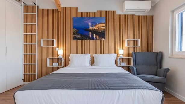 A bedroom with a large bed, wooden panel wall, a painting of a coastal scene, two bedside tables, a chair, and a window. - Noah's Ark by LovelyStay (Cascais)