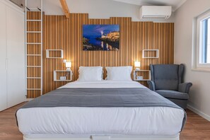 Ein Schlafzimmer mit einem großen Bett, einer Holzwand, einem Gemälde einer Küstenszene, zwei Nachttischen, einem Sessel und einem Fenster.