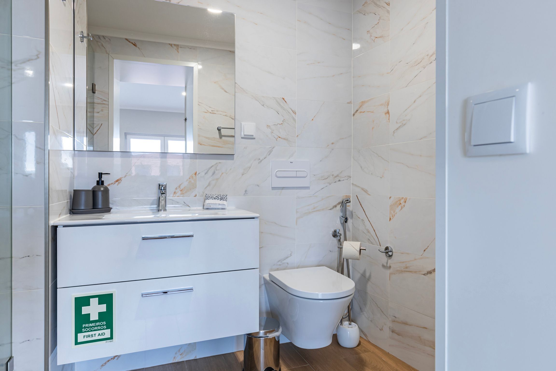Un baño moderno con pared de mármol, un lavamanos blanco con espejo arriba, un inodoro y un cartel de primeros auxilios en el mueble.