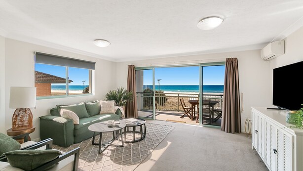A spacious living room with a green sofa, a coffee table, and a TV. It features a large sliding glass door leading to a balcony with a view of the beach and ocean. - Golden Sea 12 - Absolute Beachfront Bilinga - Min. 3 Night Stays! (Bilinga)