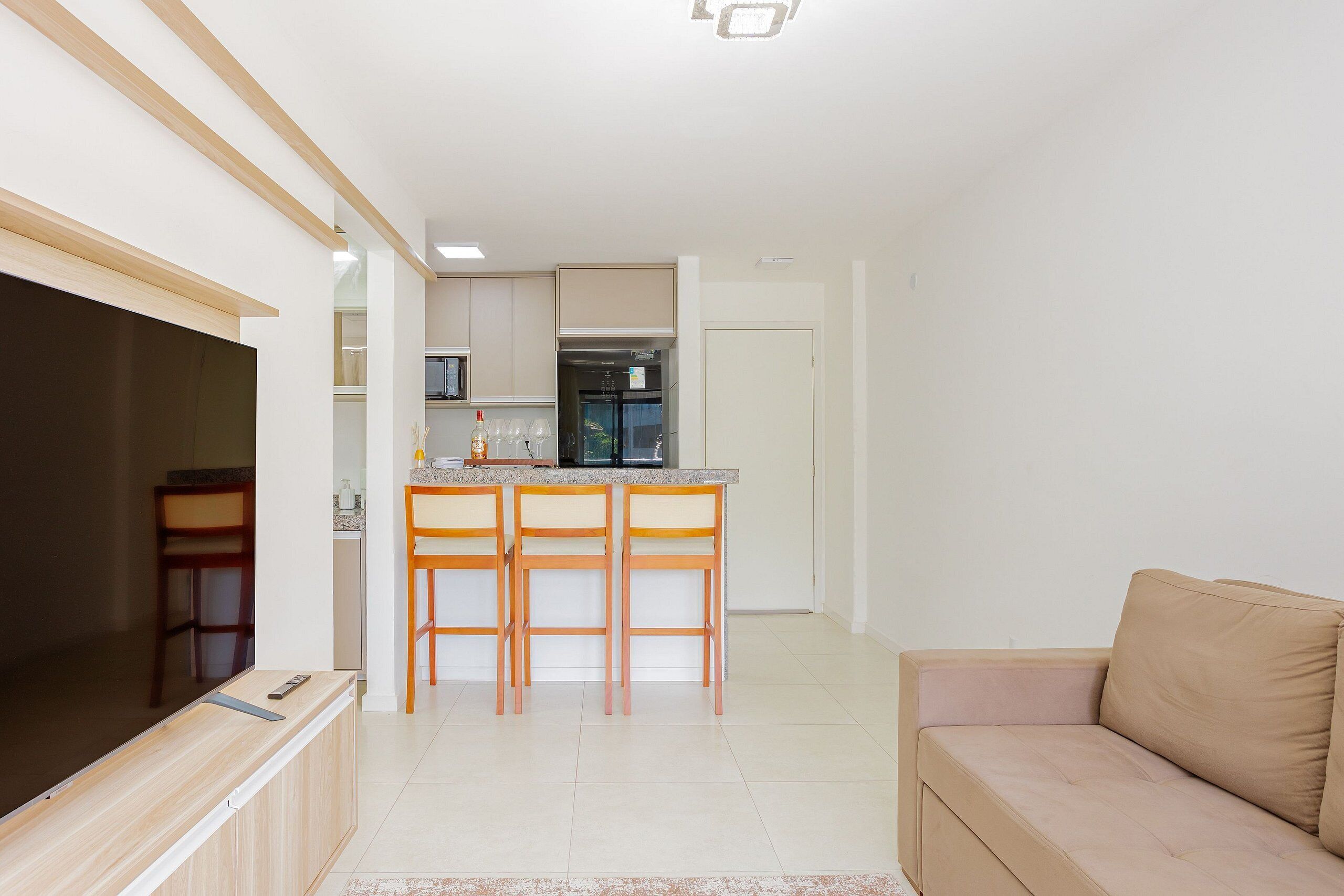A modern living room with a beige sofa, a TV on a wooden stand, and a kitchen area with a dining set.