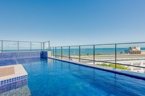 Uma piscina infinita no terraço, com águas azul-claras, guarda-corpo de vidro e vista para o oceano e edifícios ao longe.