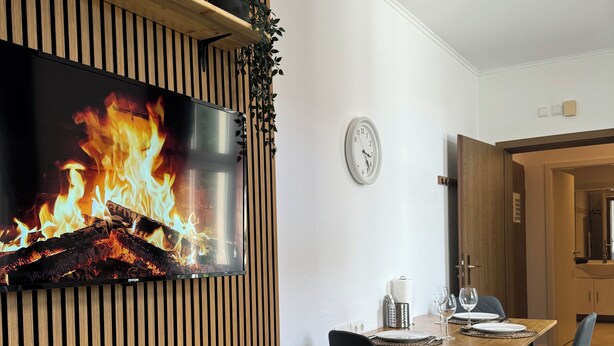 A modern dining area with a wooden slatted wall, a flat-screen TV displaying a fire, a dining table set with chairs, and a clock on the wall. - Zentral & Ruhig im Ortskern von Bad Rothenfelde (Bad Rothenfelde)