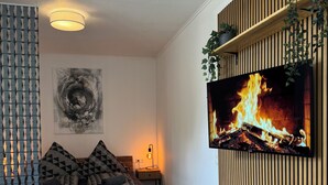 A modern living room with a wooden floor, a sofa, a coffee table with a remote control, a TV displaying a fireplace, and a wall-mounted shelf with plants. - Zentral & Ruhig im Ortskern von Bad Rothenfelde (Bad Rothenfelde)